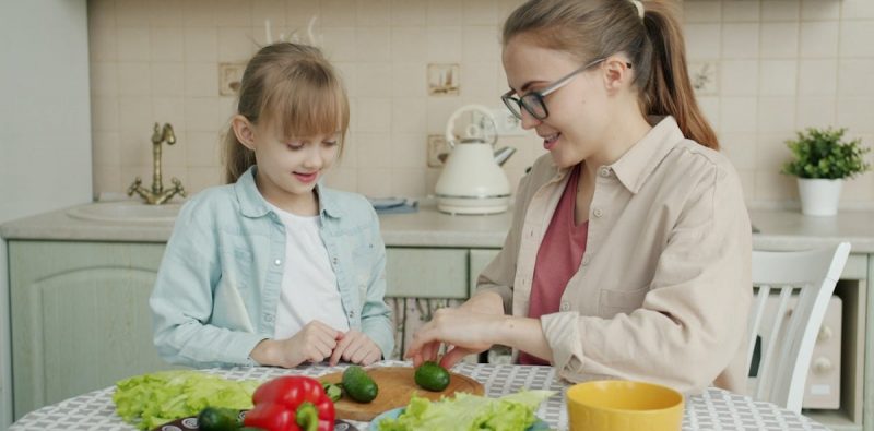 Young woman is cooking salad and feeding cute little daughter with cucumber slices in kitchen, family is talking and having good time together.